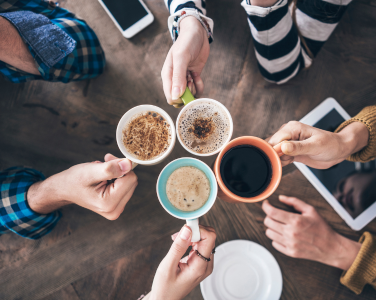 Hands holding colorful mugs of coffee over a wooden table.