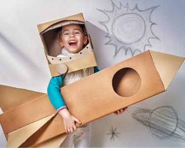 Child in a cardboard astronaut costume holding a cardboard rocket.