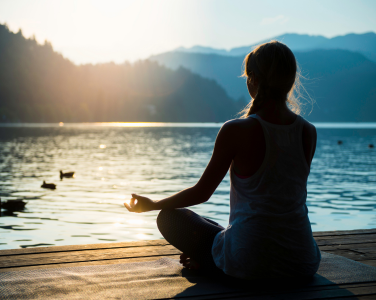 Person meditating on a dock by a lake with mountains and sunset in the background.