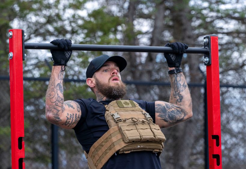 A person wearing a tactical vest and gloves performs a pull-up on an outdoor pull-up bar structure. The person has visible arm tattoos and is using a red metal pull-up frame. Trees and a fence appear 