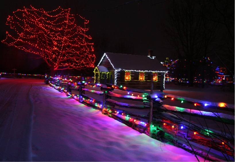 Winter holiday lights decorate a snowy cabin, fence, and tree.