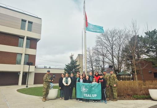 Group of civilians and CAF members in front of raised up Teal Up Flag
