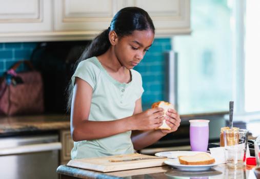 A multiracial 10 year old girl at home, sitting at the kitchen counter, about to eat a peanut butter sandwich that she just finished making