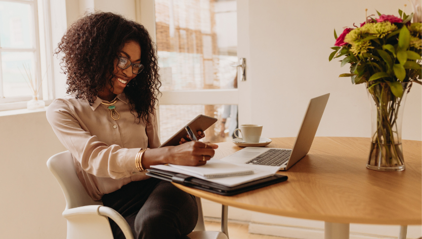 a smiling woman sitting at a table with a laptop and coffee in front of her. With a tablet in one hand, she is making notes on a pad of paper beside the laptop