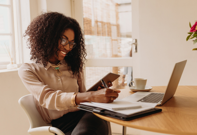 Focused businesswoman in a crisp white room, seated cross-legged at a round table with a laptop open, tablet in her left hand, pen in her right, and a coffee beside her