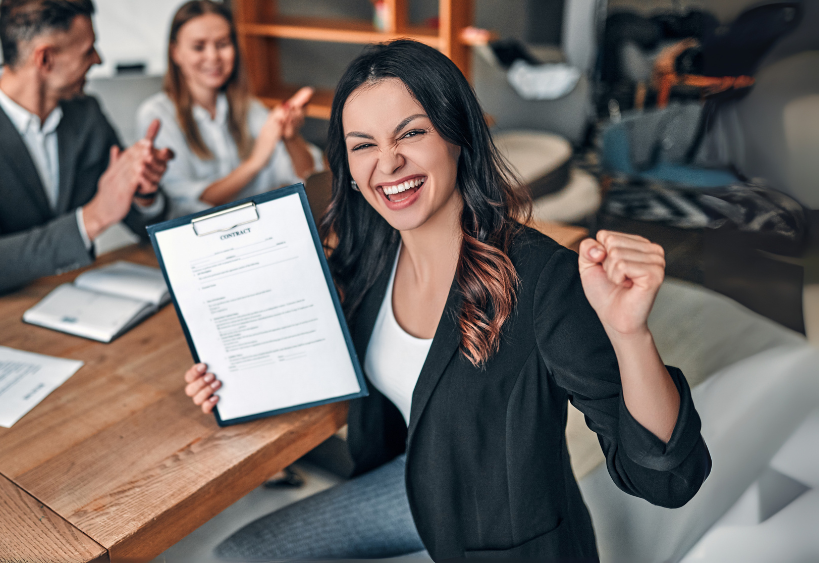 Woman in a black blazer celebrates a signed contract, raising a clipboard triumphantly as colleagues applaud in a bright office—marking a moment of success and teamwork