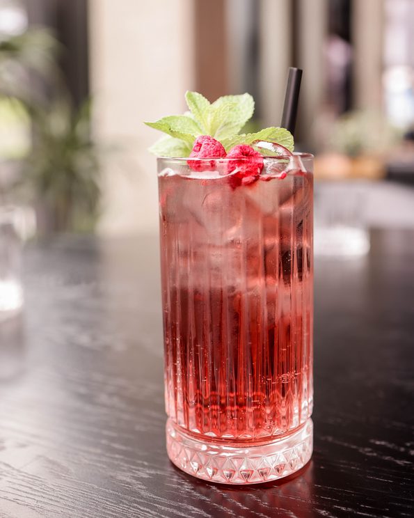 pink mocktail in a tall glass tumbler with raspberry and mint garnish sitting on a black tabletop