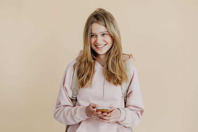 A cheerful teenager with blonde hair wearing a pink sweater and using a smartphone indoors.