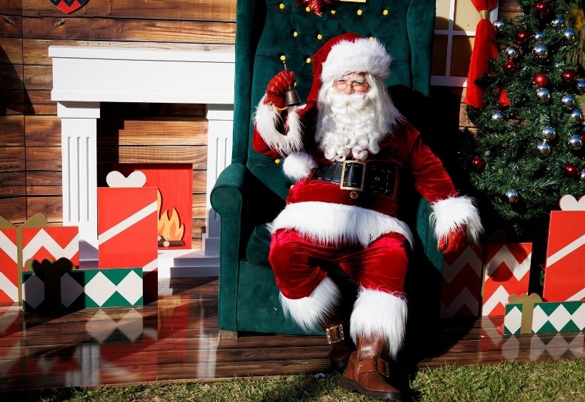Santa Clause is sitting in a big chair ringing a bell