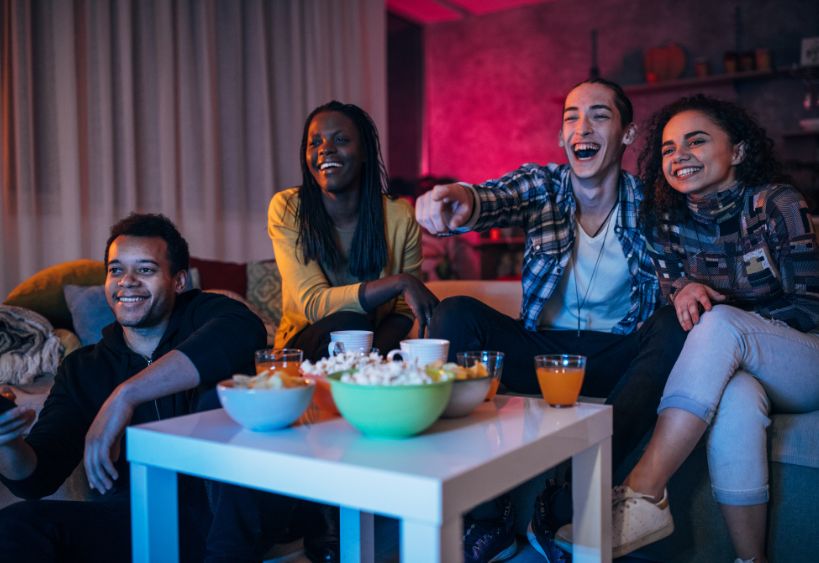 A group of friends is gathered in a living room, watching a screen while sharing popcorn and snacks.