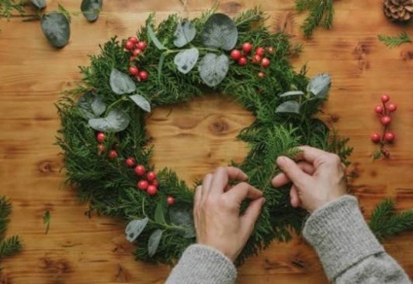 A festive holiday wreath made of fresh evergreen branches, eucalyptus, and red berries is being carefully crafted on a wooden table.