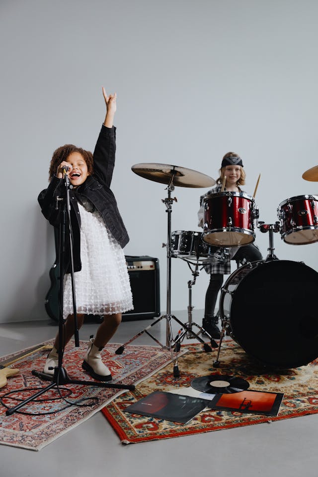 Two young kids perform energetically in a music band with drums and a microphone.