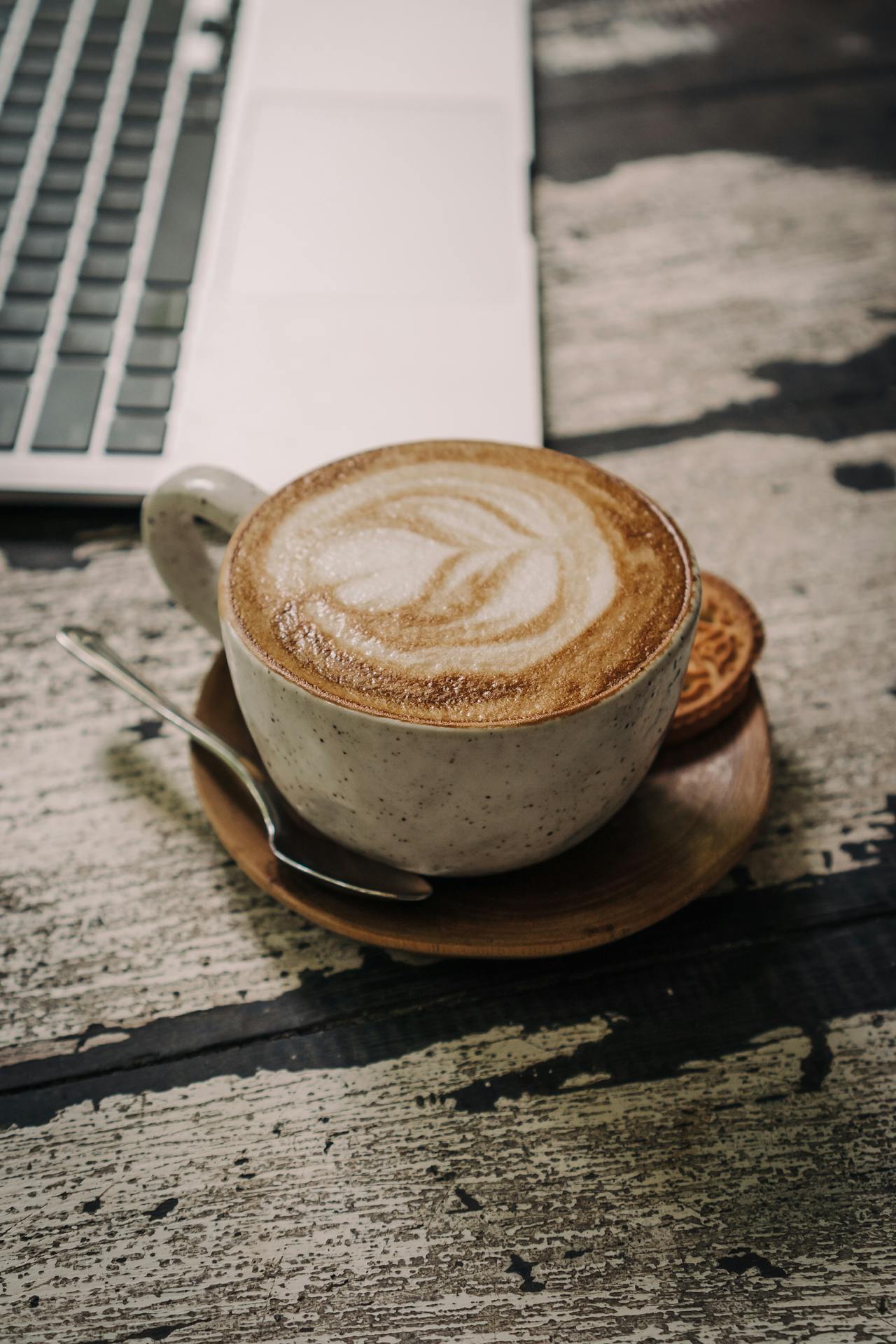 A close-up of a cappuccino cup and saucer on a rustic table next to a laptop.