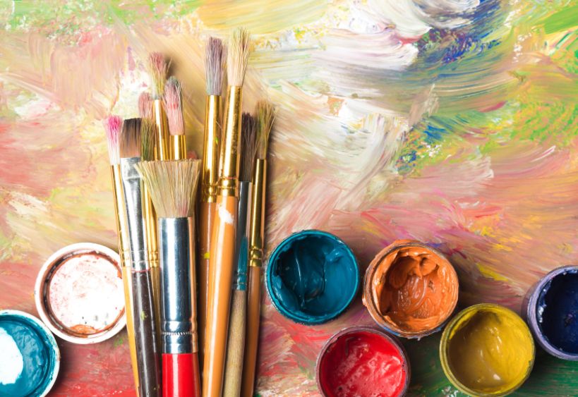 Paint brushes and pots of paint laid on an abstract colourful painting.