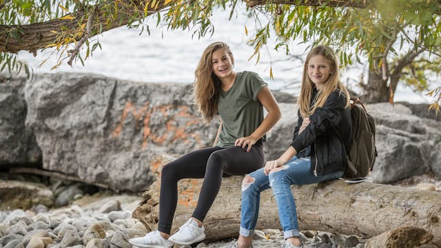 two teens sitting on a rock along a shoreline with a tree hanging down
