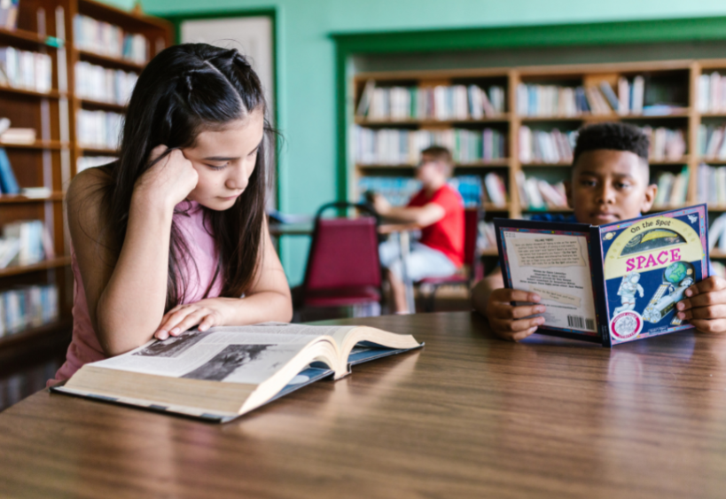 two children reading in a library, sitting at a table