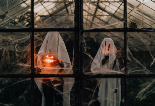 2 people dressed as ghosts standing in a window - one holding a jack-o-latern