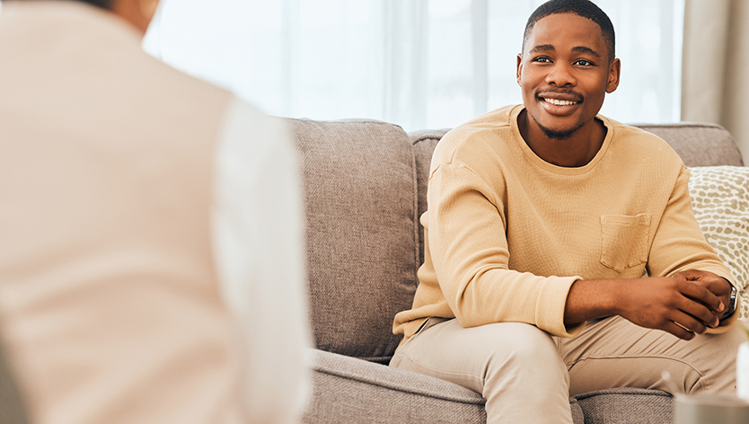 Man sitting on couch smiling, and back of person talking to man on couch