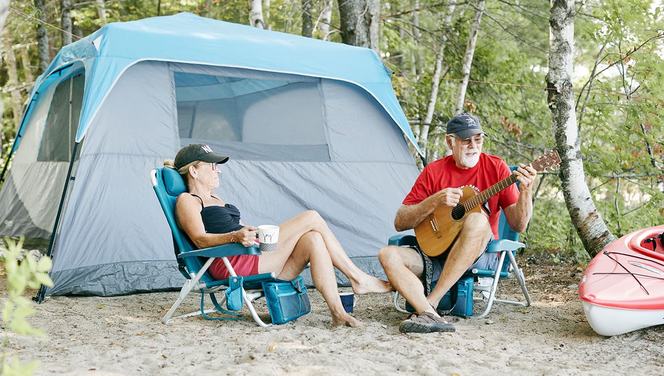 Couple camping sitting in front of tent. Man playing guitar, woman drinking coffee