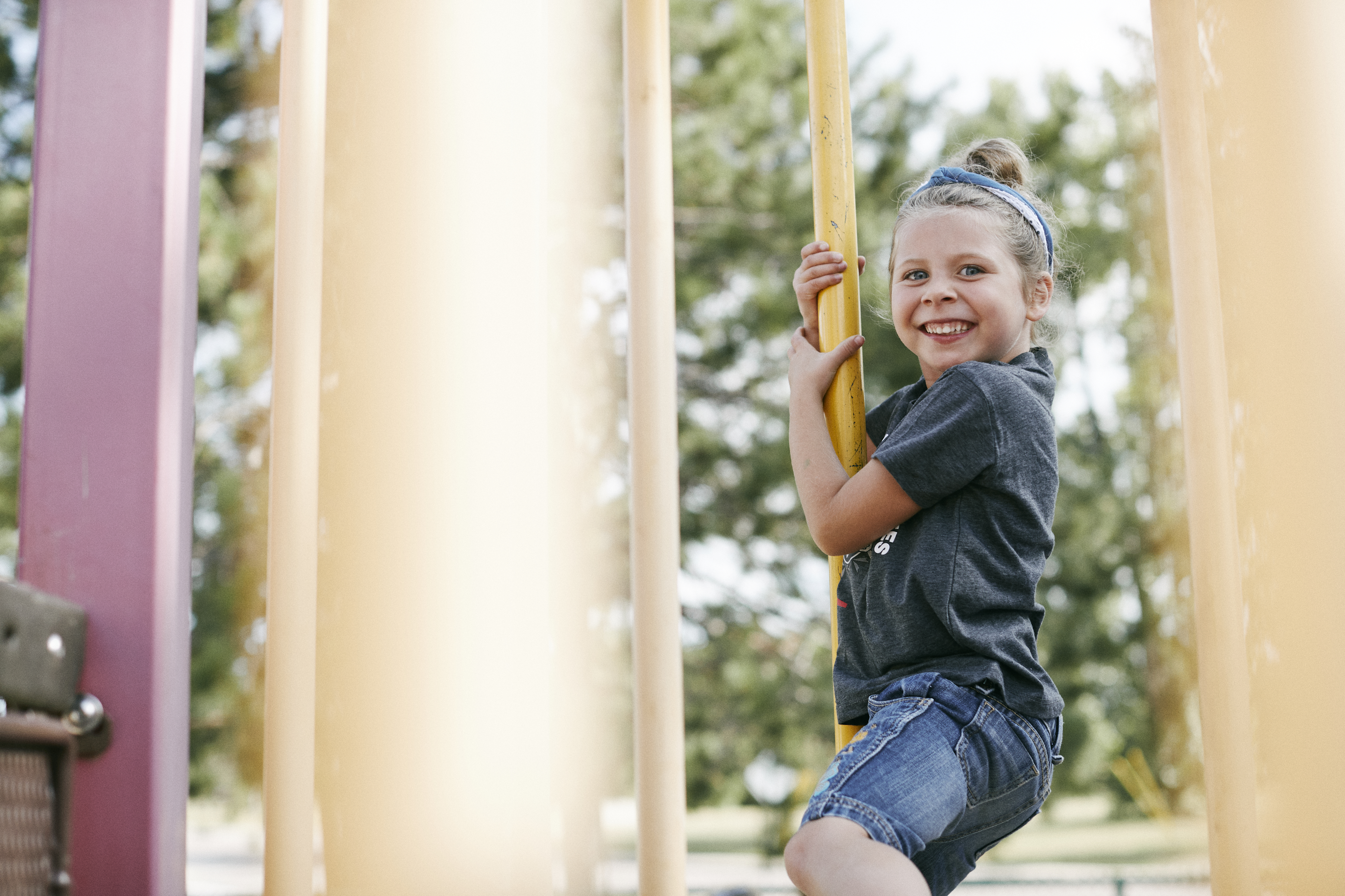 Image of a school aged girl with blonde hair playing on a play structure smiling at the camera.
