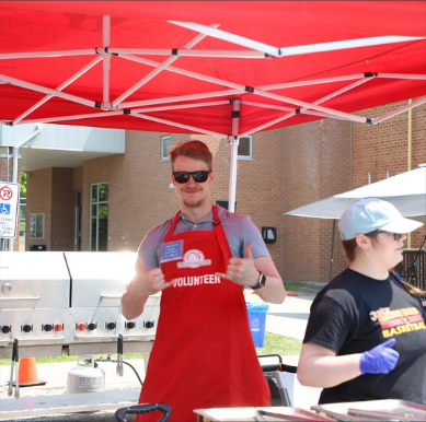Two volunteers stand outdoors under a red canopy at a community event. One volunteer, wearing a red volunteer apron and sunglasses, smiles and gives a thumbs-up while another volunteer works nearby.