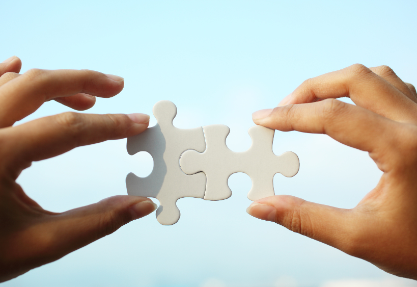 Two hands poised to connect puzzle pieces against a clear blue sky—capturing the moment where collaboration meets clarity