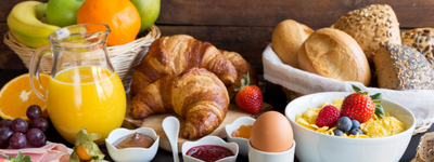 A breakfast spread with orange juice, croissants, bread rolls, fruit, yogurt with berries, jam, and a boiled egg on a wooden table.