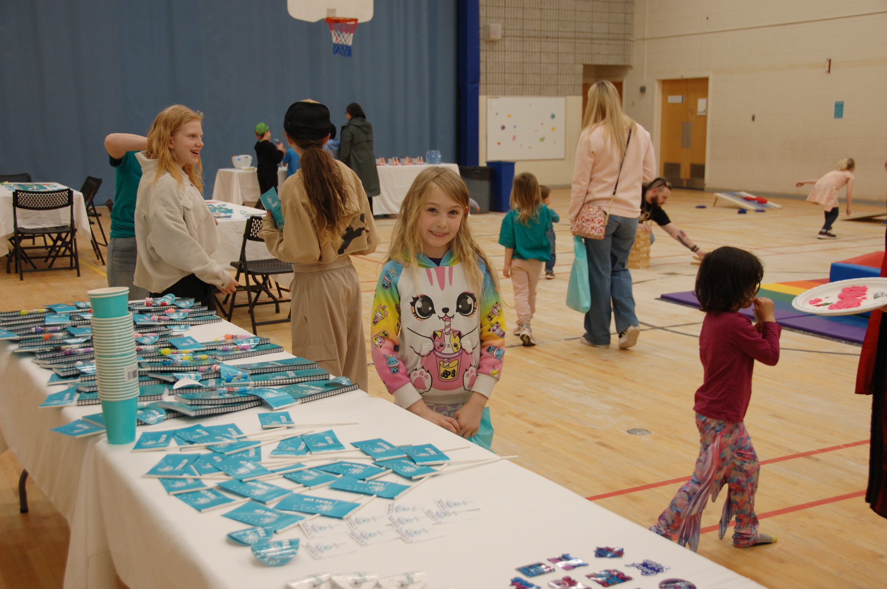 Children and families participate in a community event inside a gymnasium, with kids playing, doing crafts, and visiting a table with teal-themed items and giveaways.