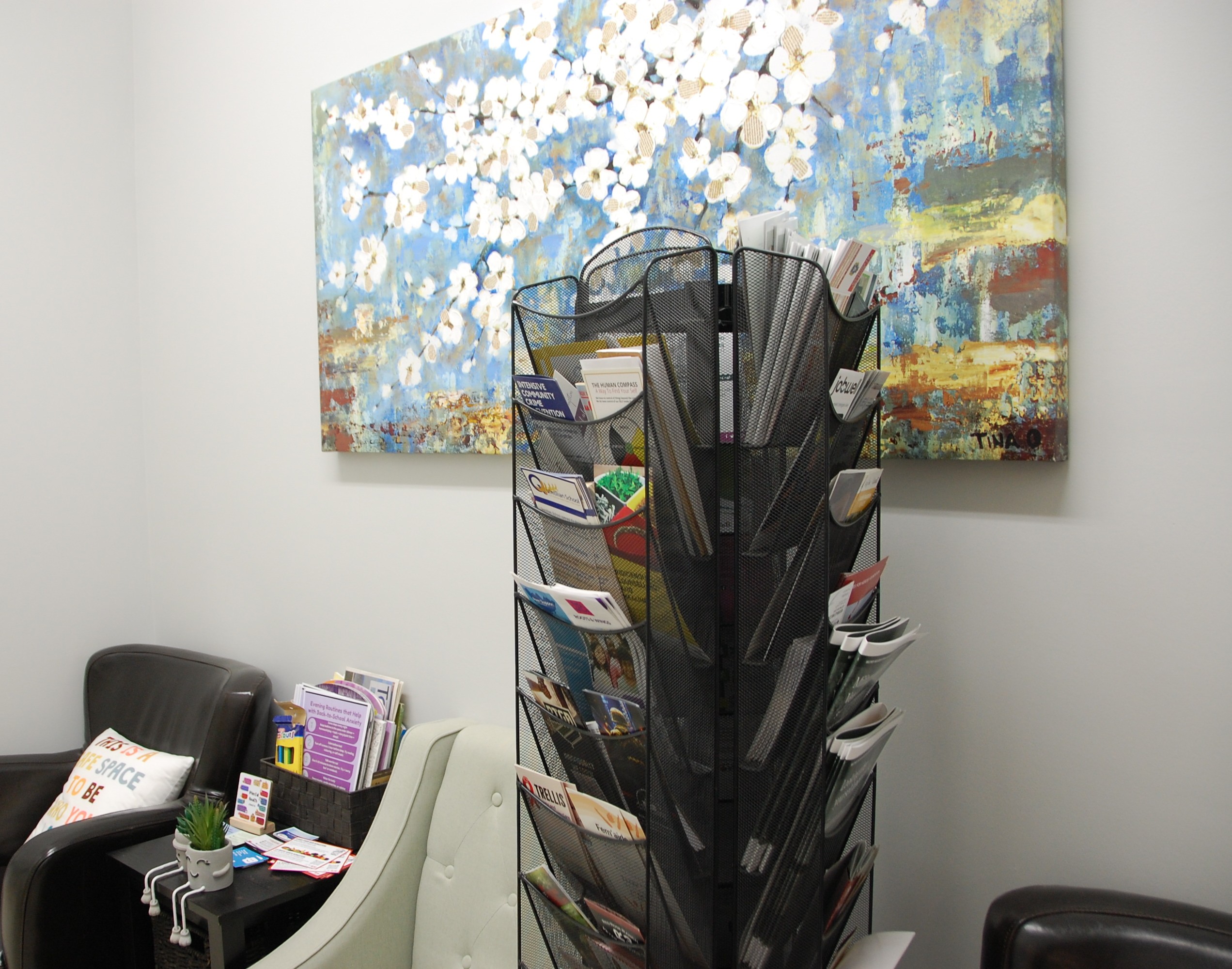 Bright and welcoming mental health waiting area with comfortable armchairs, a small side table with brochures, a rotating literature rack filled with community resources, and calming floral wall art.