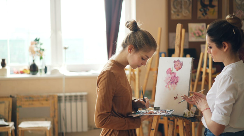 Two women are painting flowers on a canvas that is on an easel